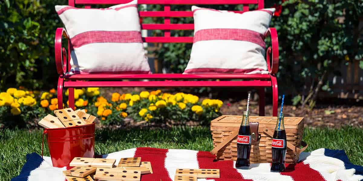 A red garden bench with cushions behind a picnic blanket set up with a game and drinks in a backyard.